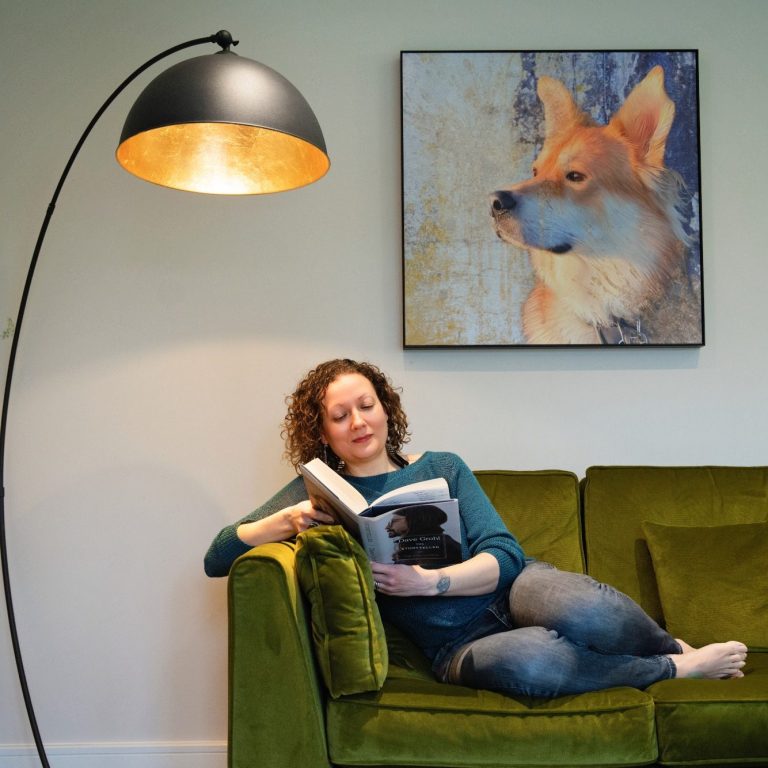 A woman reading on a green sofa, with a lamp and a dog painting on the wall.