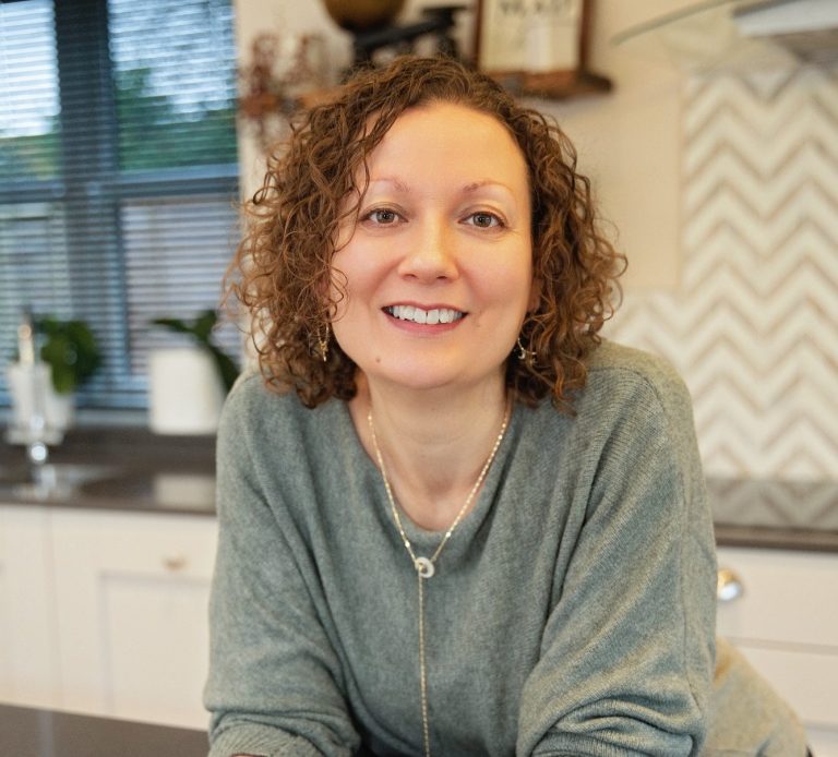 Smiling woman with curly hair in a kitchen, wearing a grey green sweater.
